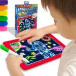 Niño usando pizarra digital roja con luz LED para dibujar un astronauta, incluye marcadores de colores y empaque del producto con diseño espacial.