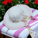Sombrero de paja blanca de ala ancha con borde rosado y lazo de tela con estampado de rosas, sobre una superficie de rayas blancas y rosadas.