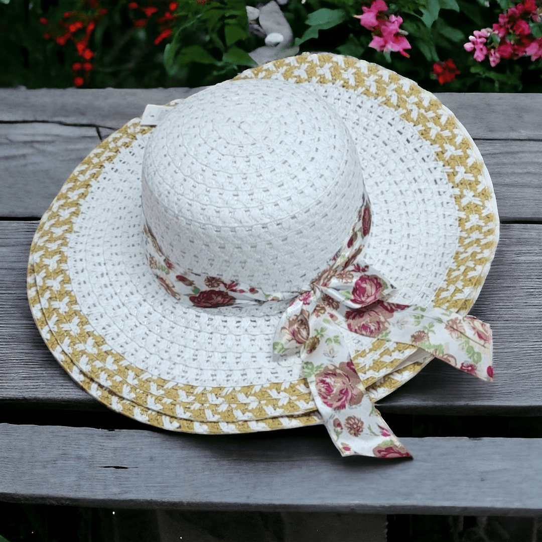 Sombrero de ala grande en fibra blanca con borde dorado, cinta estampada de rosas y lazo lateral, sobre superficie de madera.