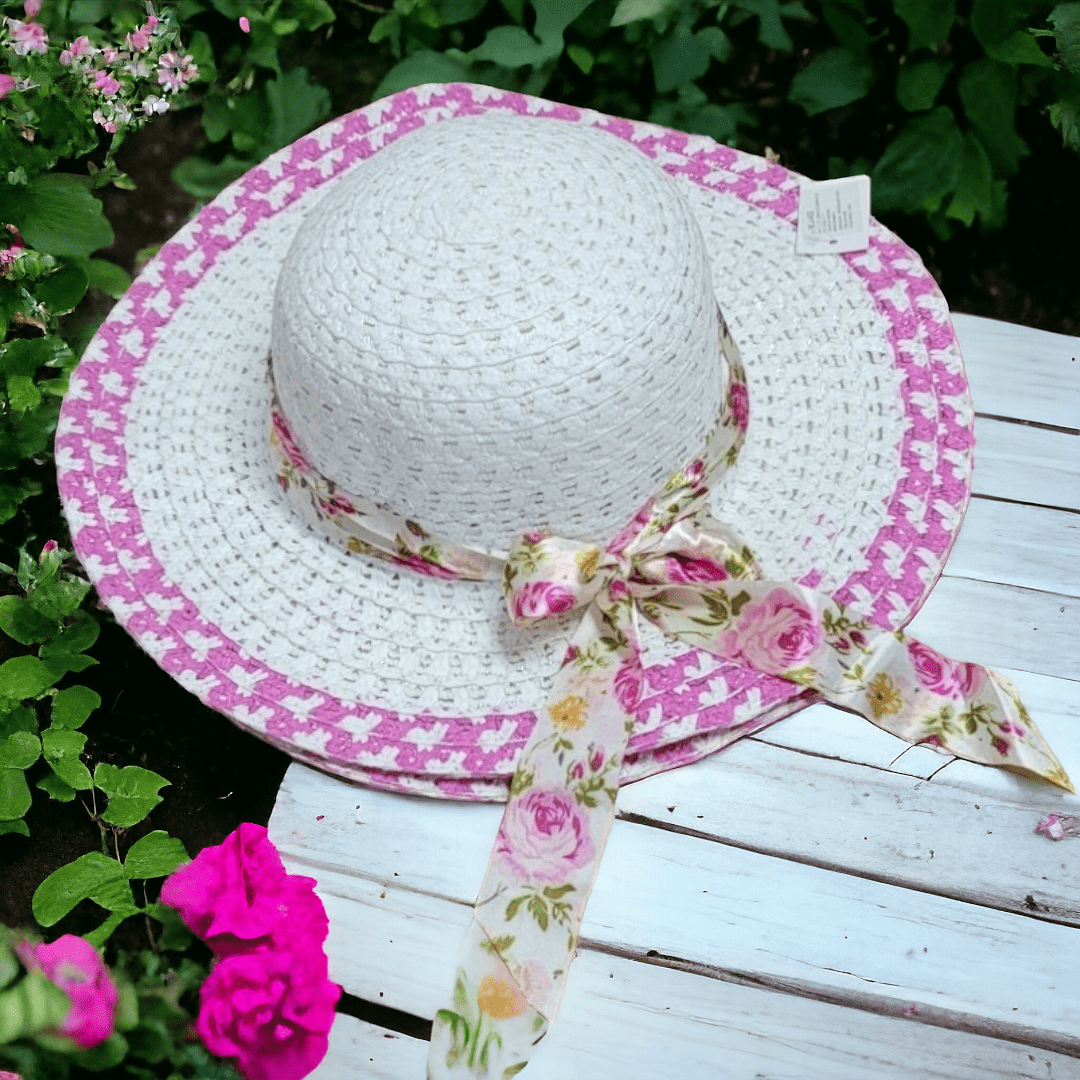Sombrero de paja blanca de ala ancha con borde rosa y lazo de tela con estampado floral, sobre mesa de madera blanca en un jardín.