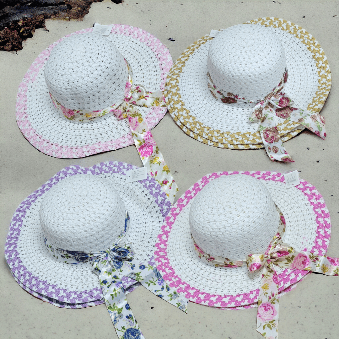 Cuatro sombreros de ala ancha blancos de paja sobre arena, cada uno con un lazo de tela estampado diferente en tonos rosa, amarillo y azul.
