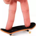 Dedo índice y medio presionan una mini patineta de madera con lija negra y ruedas blancas sobre fondo blanco.