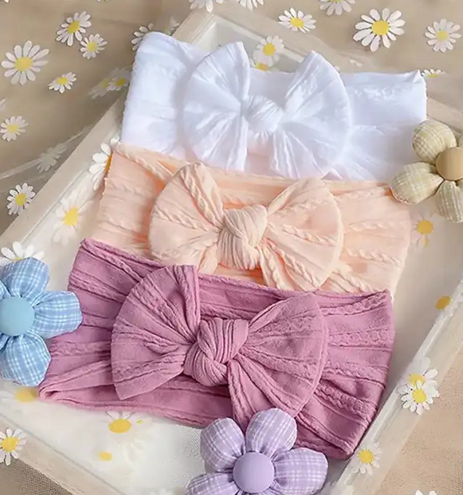 Tres diademas de nailon para bebé con lazos de textura trenzada en colores blanco, durazno y malva, dispuestas sobre una bandeja decorada.