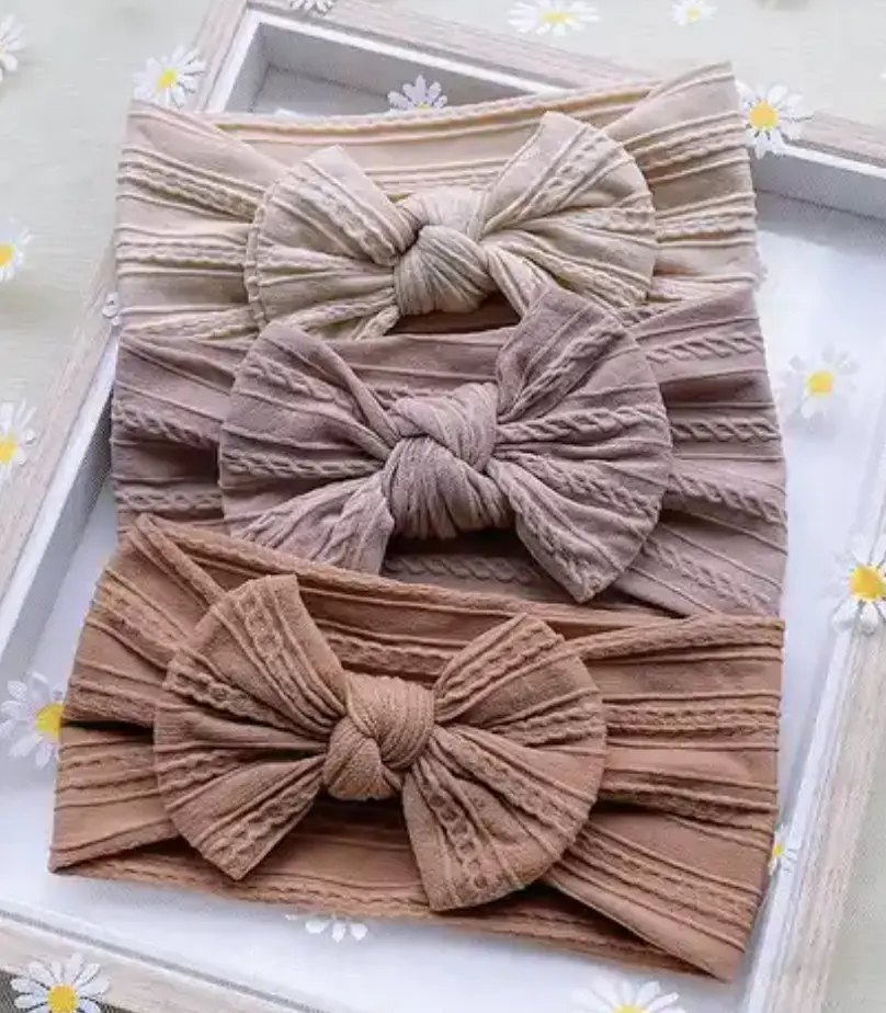 Tres diademas de nailon para bebé con lazos grandes y textura acanalada en tonos beige, rosa viejo y marrón, sobre una bandeja con margaritas.