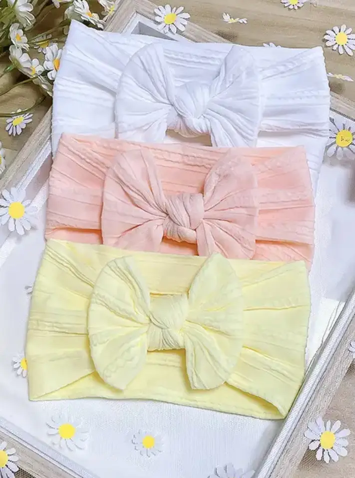 Set de tres diademas de nailon elástico para bebé con lazo suave en colores blanco, rosa pastel y amarillo, dispuestas horizontalmente sobre bandeja blanca.