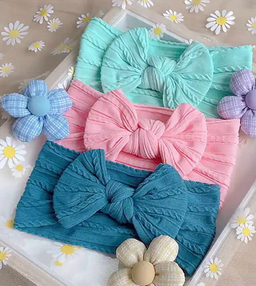 Tres diademas de nailon para bebé con lazos de textura trenzada en colores menta, rosa y azul petróleo sobre una bandeja con motivos florales.
