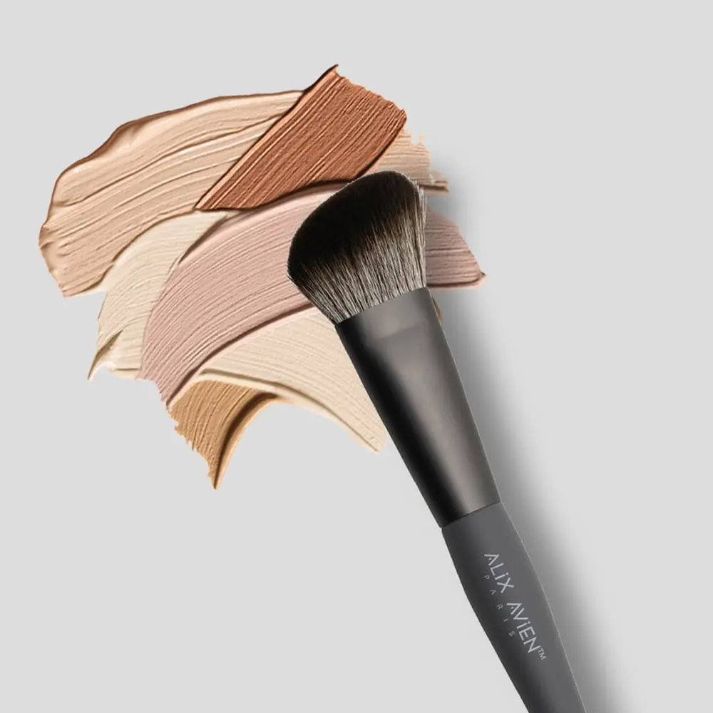 Brocha angulada Alix Avien de mango negro sobre trazos de maquillaje líquido en diversos tonos de piel para contorno e iluminador.