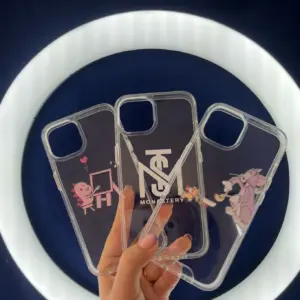 Set de tres forros transparentes para iPhone con diseños variados de dinosaurio, logotipo Monastery y Tom y Jerry, sostenidos por una mano.