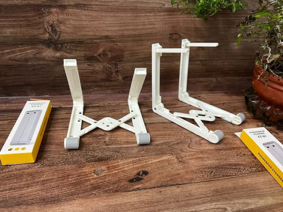 Set x2 soportes ajustables blancos de plástico para celular y tablet, con diseño plegable, brazos extensibles y cajas de empaque sobre superficie de madera.