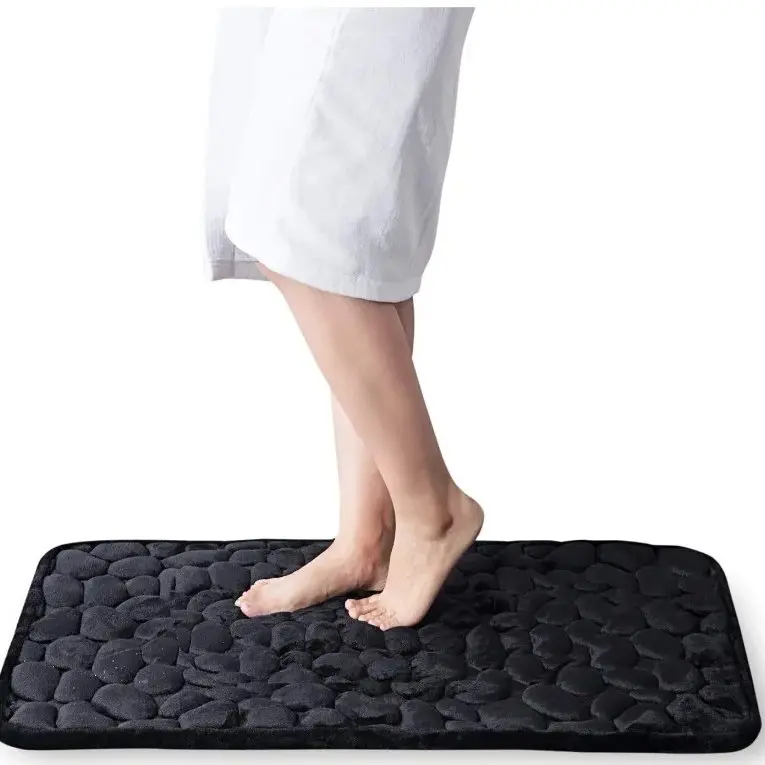 Tapete de baño negro con relieve de piedras en espuma viscoelástica, usado por una persona descalza para mostrar su textura y confort.