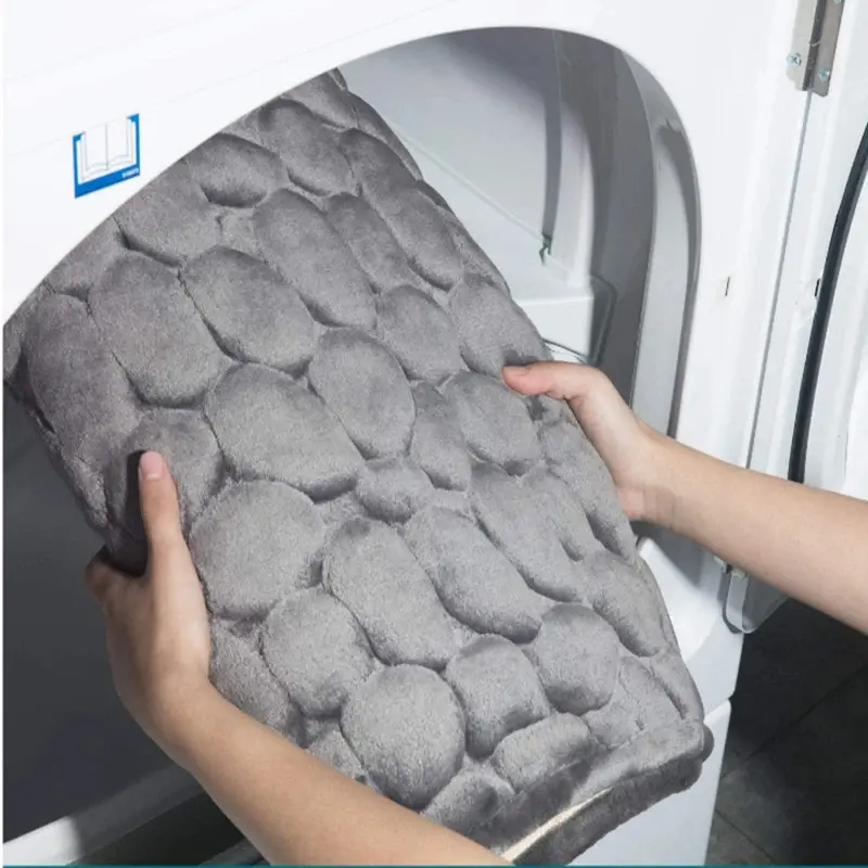 Tapete de baño gris con textura de piedras en relieve, siendo colocado en una lavadora blanca para resaltar su facilidad de lavado.