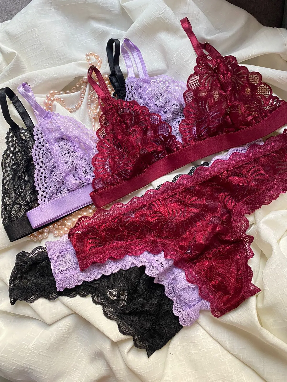 Set de ropa interior para mujer en encaje suave, color vino tinto, lila y negro, brasier triángulo y panty delicados sobre fondo blanco.