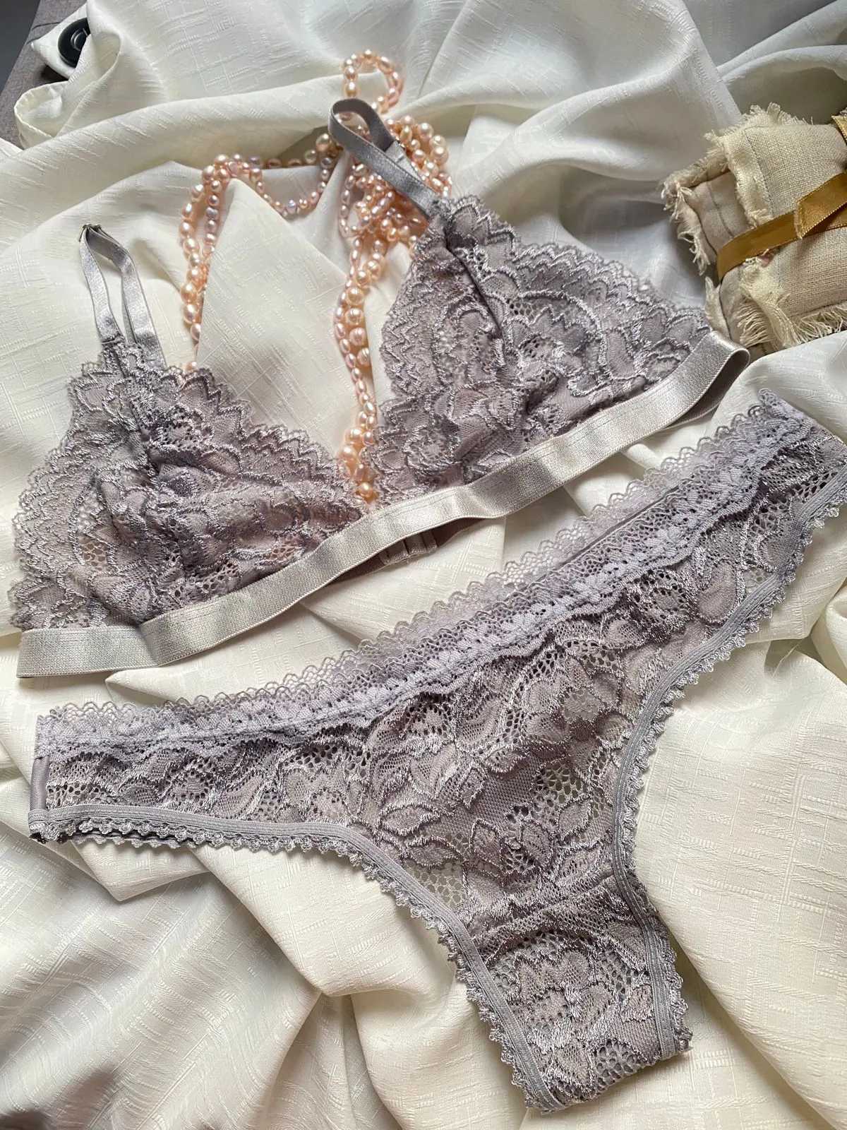 Conjunto de lencería en encaje color malva con sujetador de tirantes finos y panty a juego, sobre tela blanca con un collar de perlas.