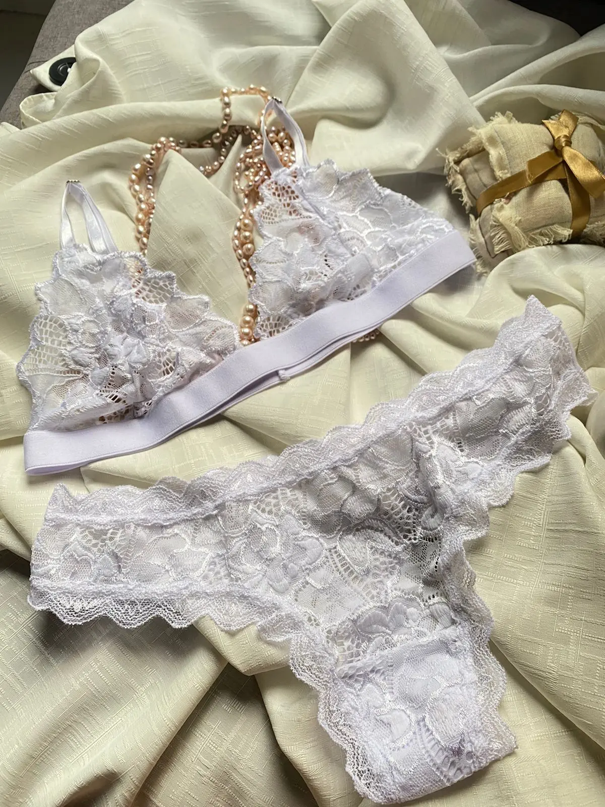 Set de lencería blanca para mujer con sujetador de encaje y banda elástica, acompañado de bralette a juego y collar de perlas sobre tela beige.