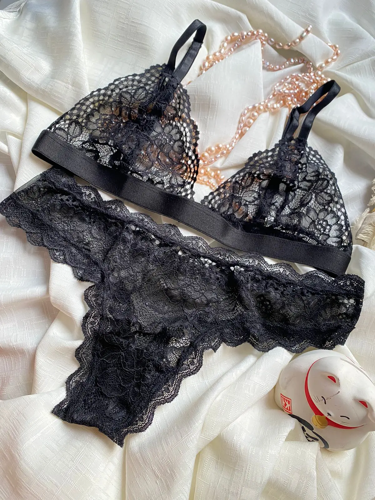 Conjunto de lencería negra en encaje suave, compuesto por bralette de tirantes ajustables y tanga escotada, sobre fondo blanco textil con collar de perlas rosadas.