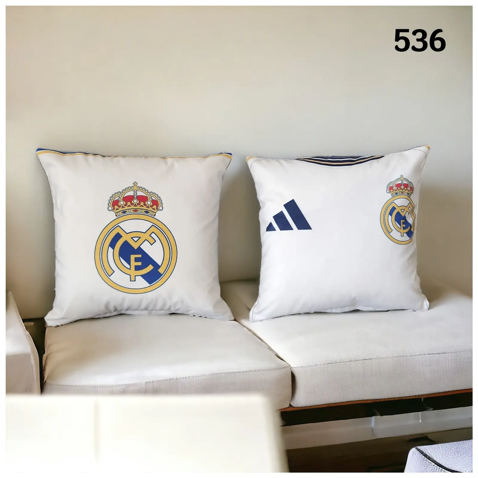 Set de dos fundas de cojín blancas decorativas con el escudo del Real Madrid y logo de Adidas, dispuestas sobre un sofá blanco.