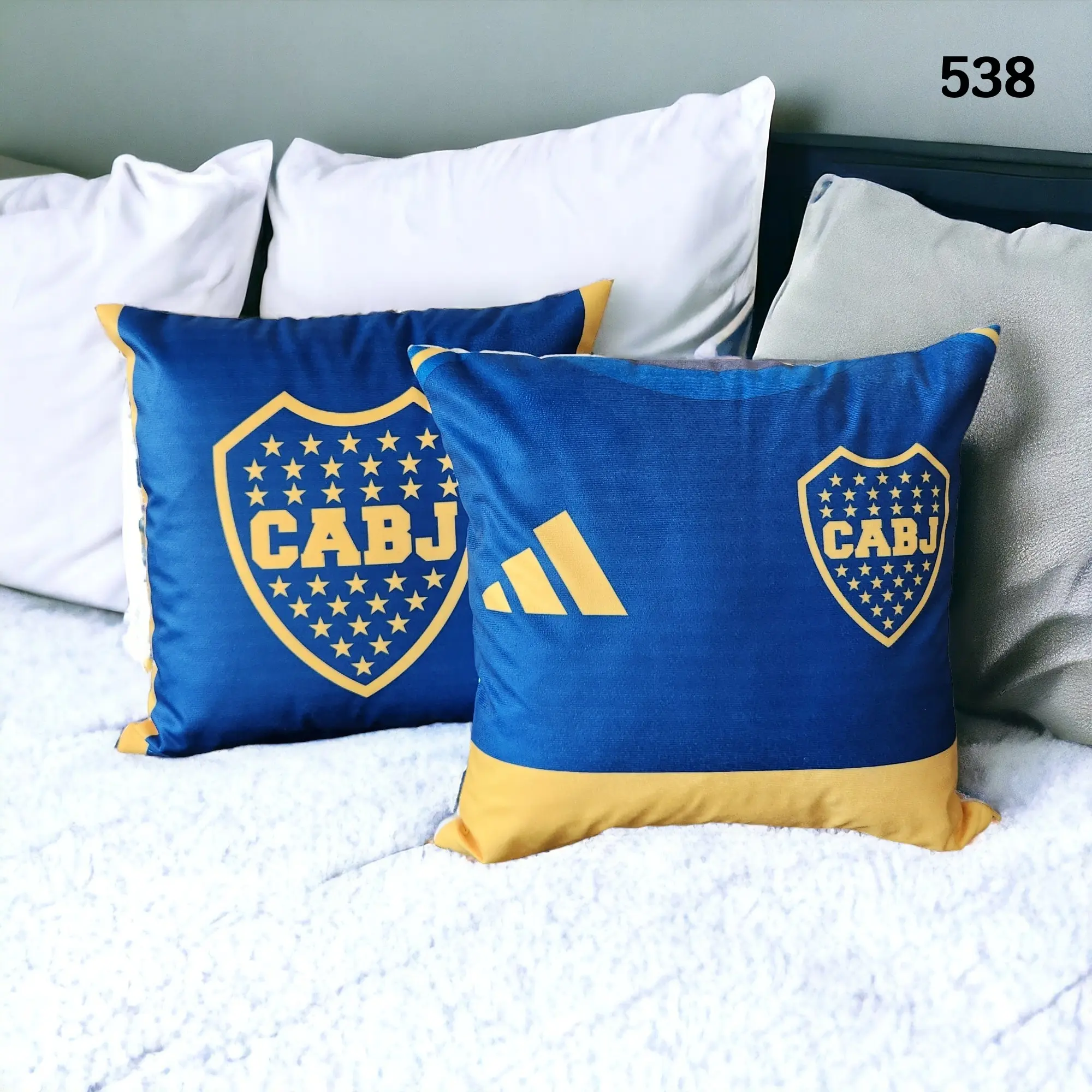 Dos fundas para cojines azules y amarillas del club Boca Juniors con escudos y logos, sobre una cama con ropa blanca texturizada.