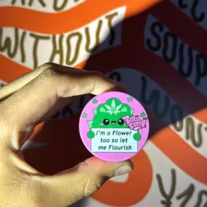 Moledor de hierbas rosa de tres pisos con diseño ilustrado de cactus verde y frase en la tapa, sostenido frente a pared con patrones naranjas y blancos.