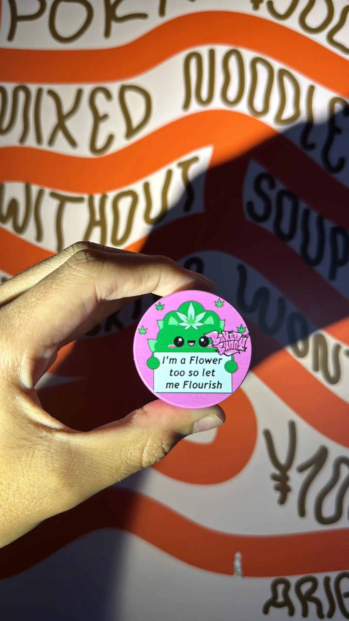Moledor de hierbas rosa de tres pisos con diseño ilustrado de cactus verde y frase en la tapa, sostenido frente a pared con patrones naranjas y blancos.