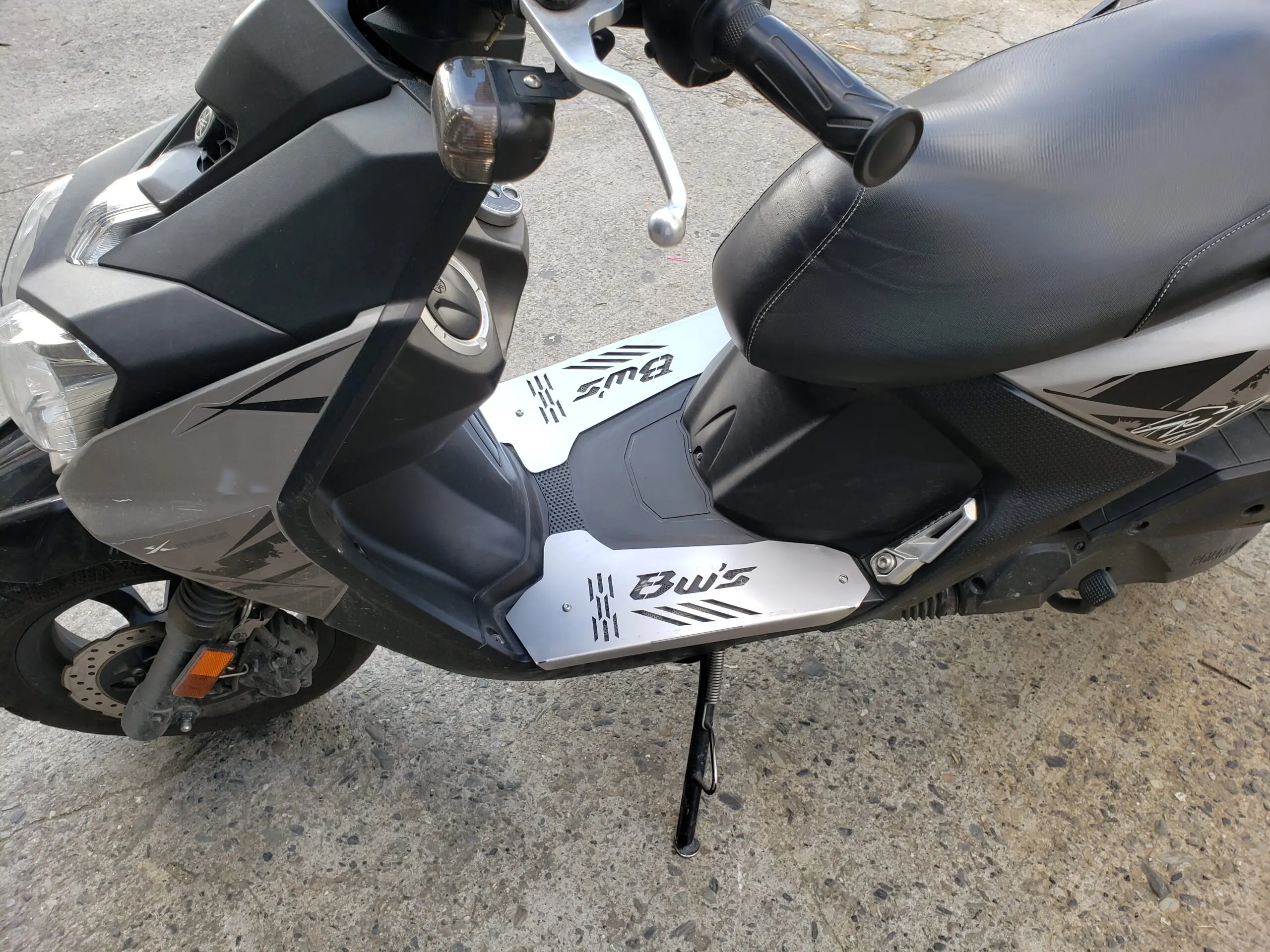 Piso protector plateado en acero inoxidable para moto BWS, instalado en la zona de los pies.
