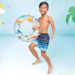 Flotador inflable circular para niños con diseño de peces coloridos sobre fondo blanco, sostenido por un niño en un entorno de piscina tropical.