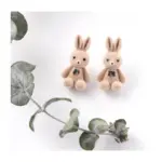 Aretes en forma de conejos beige de felpa suave con lazo negro, presentados en pareja junto a una rama de eucalipto sobre fondo blanco.