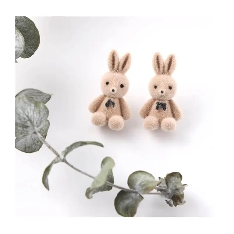 Aretes en forma de conejos beige de felpa suave con lazo negro, presentados en pareja junto a una rama de eucalipto sobre fondo blanco.