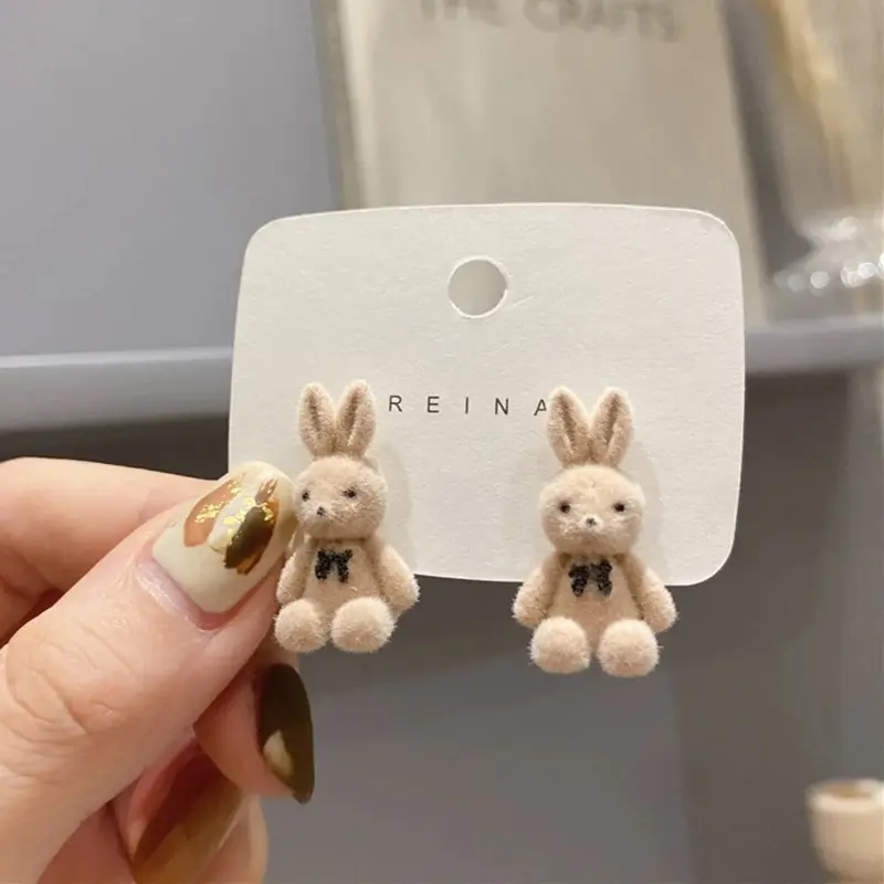 Par de aretes tipo broquel con diseño de conejitos tiernos de felpa beige y lazo negro, presentados en una tarjeta blanca.