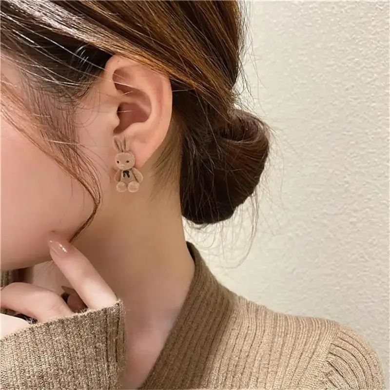 Aretes de conejo de felpa color café claro con lazo negro, puestos en la oreja de una mujer de perfil que usa suéter marrón texturizado