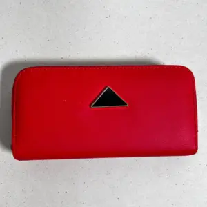Billetera roja con broche metálico triangular negro, diseño rectangular de bordes redondeados sobre fondo gris claro texturizado.