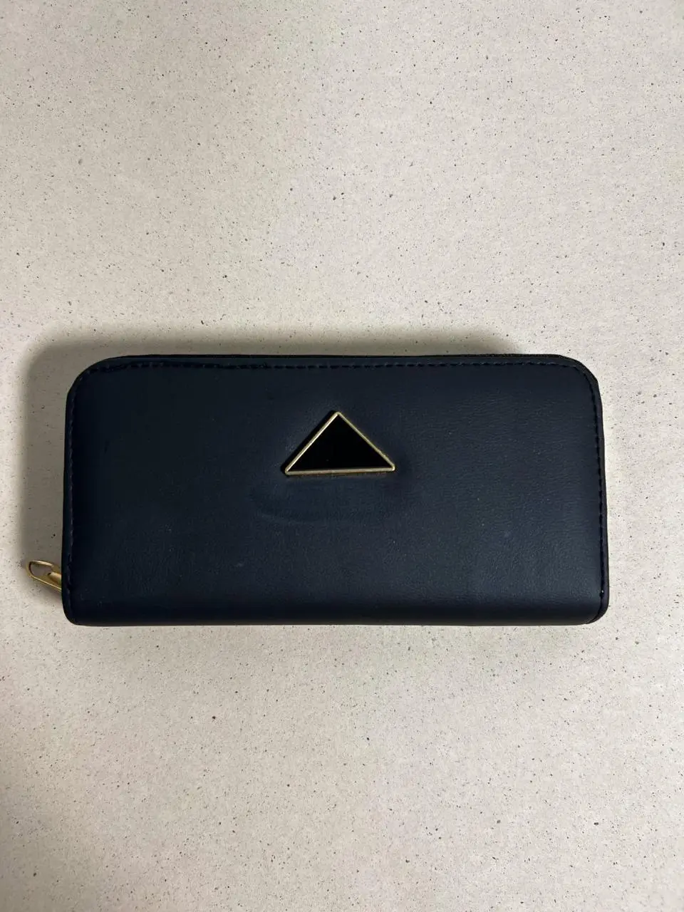 Billetera larga azul marino con broche triangular negro y borde dorado en el centro, cierre de cremallera dorado y textura lisa.