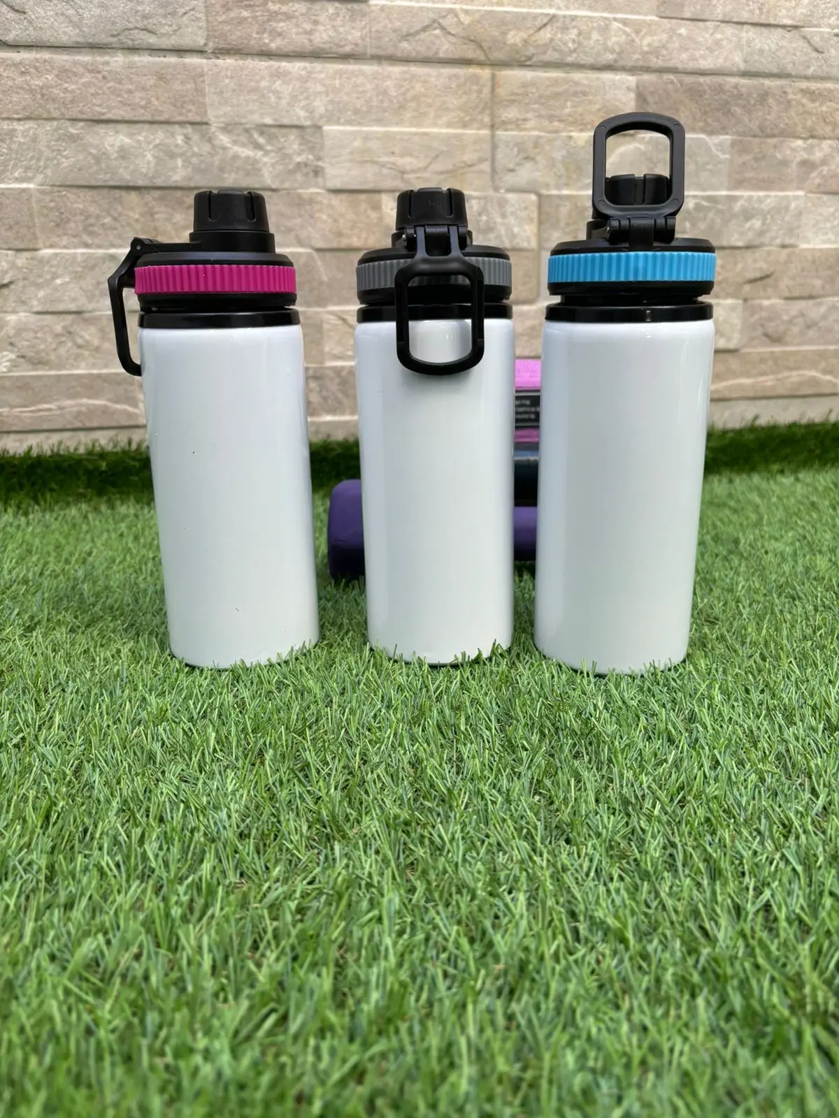 Tres botellas termo de aluminio blancas con soporte para celular en las tapas, en colores fucsia, gris y azul sobre césped artificial.