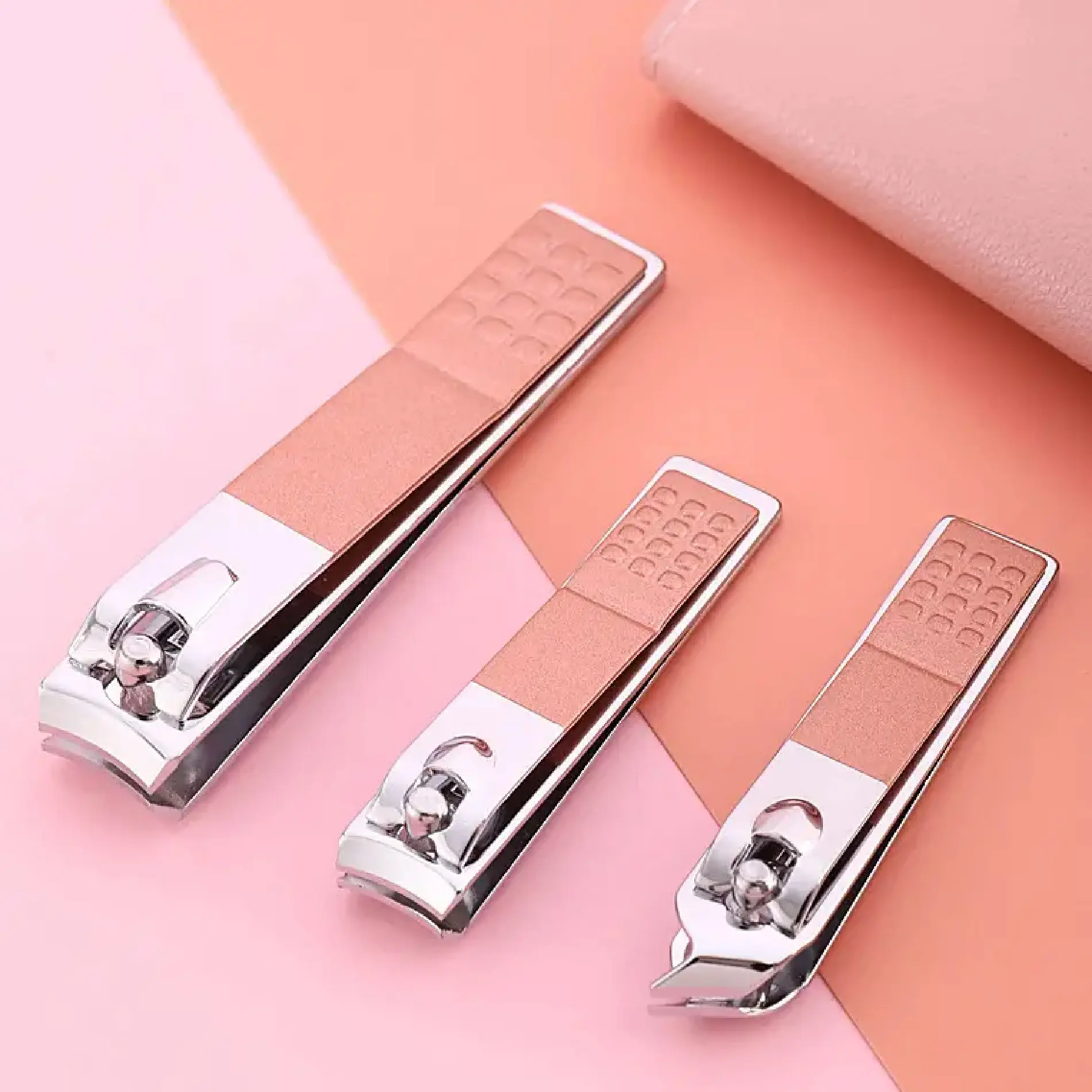 Set de tres cortaúñas metálicos de color oro rosado con mango texturizado, organizados en diagonal sobre una superficie bicolor rosa y salmón.