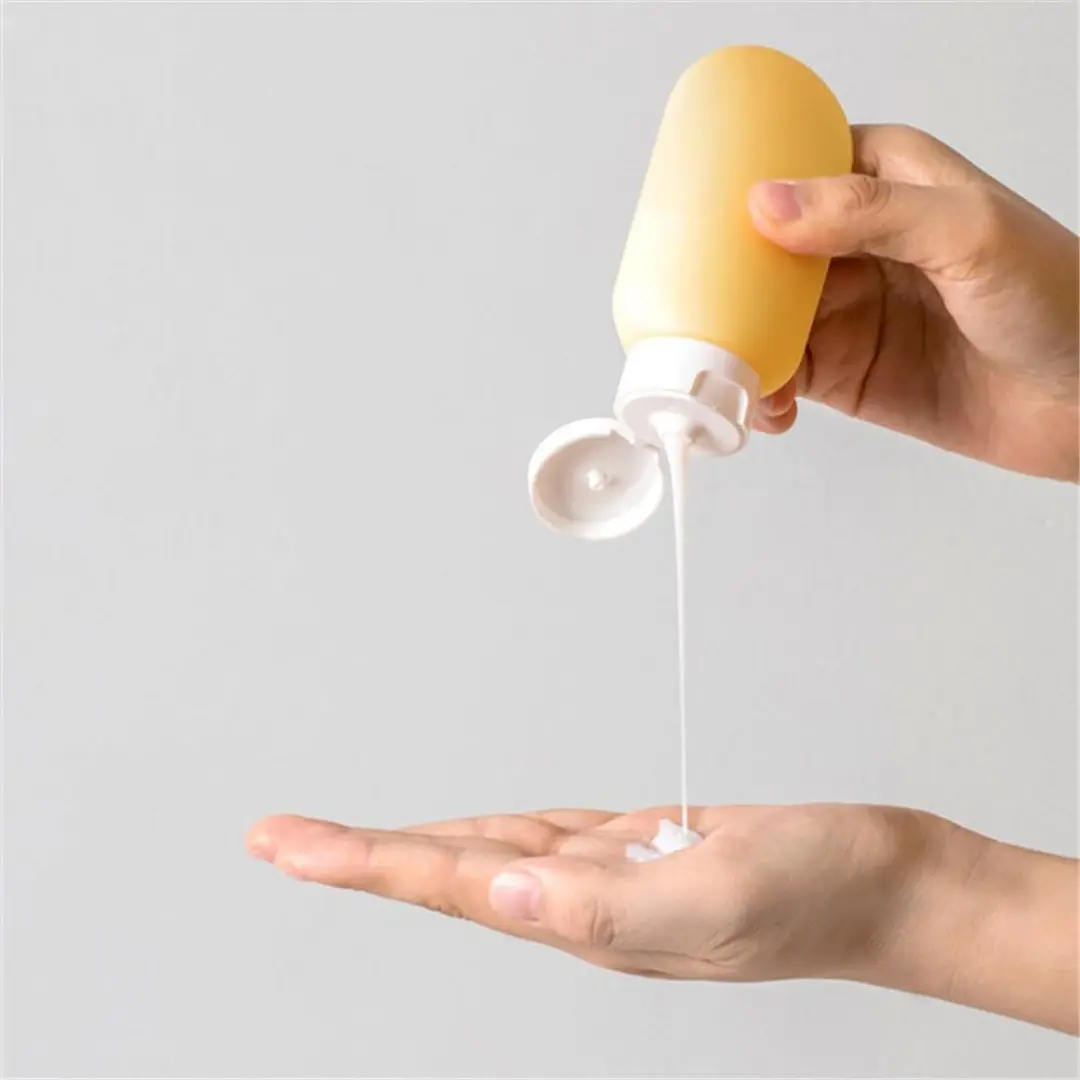 Botella de silicona amarilla exprimible con tapa dosificadora blanca, dispensando loción blanca sobre la palma de una mano para viaje.