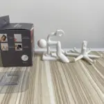 Gancho giratorio blanco de seis mordazas en caja, con base adhesiva transparente y figura humana decorativa sobre mesa de madera.