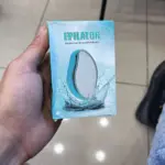 Mano sosteniendo caja azul de depilador de vidrio exfoliante con diseño ergonómico y de agua salpicando