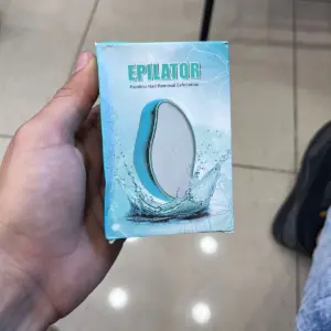Mano sosteniendo caja azul de depilador de vidrio exfoliante con diseño ergonómico y de agua salpicando