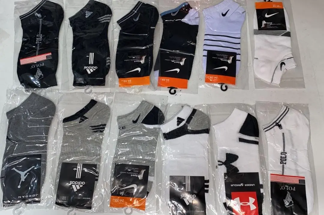 Doce pares de medias tobilleras en empaques individuales, con colores blanco, negro y gris de marcas como Nike, Adidas y Jordan.