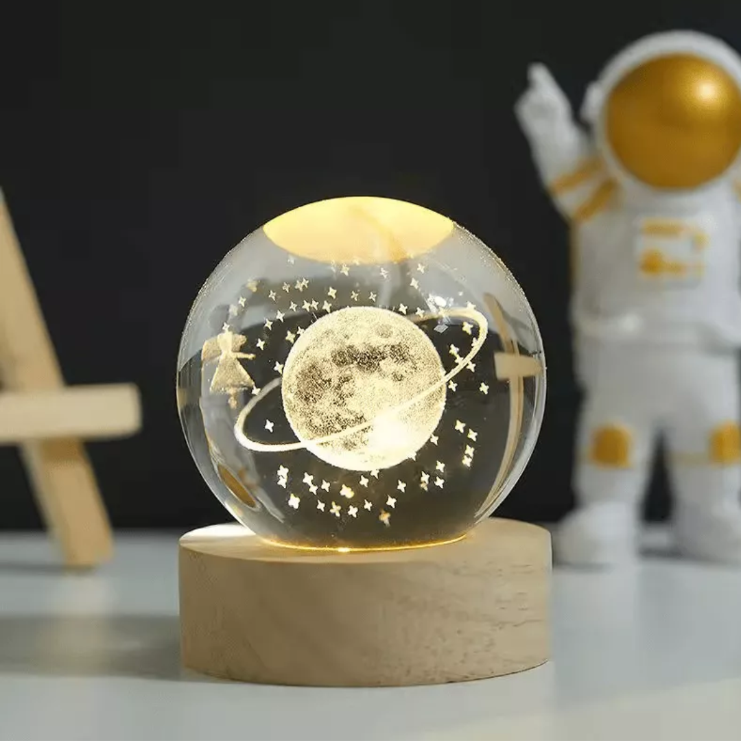 Lámpara de mesa con esfera de cristal grabada con un planeta y estrellas, sobre base de madera circular iluminada con luz cálida junto a un astronauta.