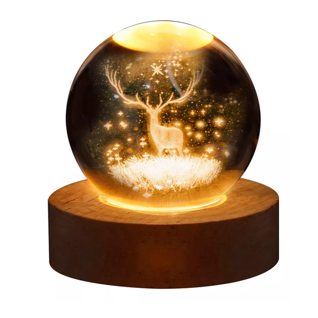 Lámpara de mesa con esfera de cristal grabada con un ciervo y estrellas, sobre base de madera circular con iluminación LED cálida conectada por USB.