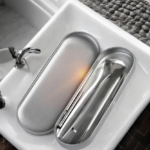 Kit limpiador de lengua de acero quirúrgico plateado con estuche metálico ovalado, mostrando dos raspadores metálicos y un palillo en su interior sobre un lavabo blanco.