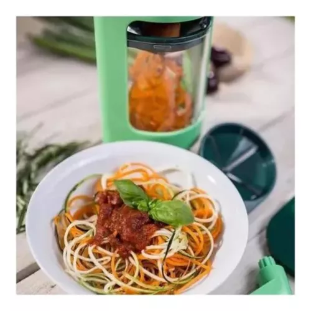 Espiralizador manual verde con dos cuchillas, procesando vegetales en su interior junto a un plato de zoodles con salsa y albahaca sobre mesa de madera.