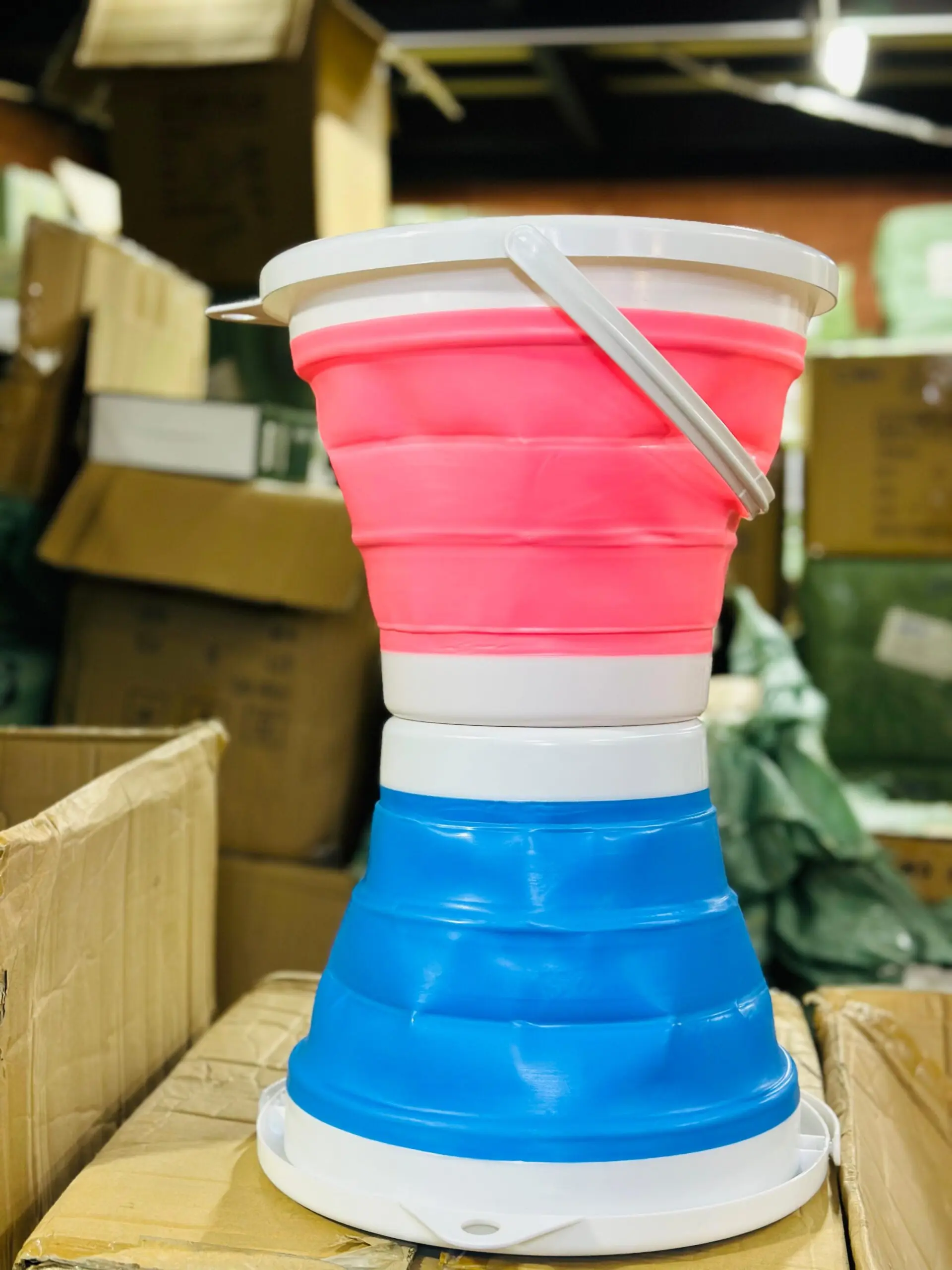 Set de dos baldes retráctiles de silicona de 10 litros, uno rosa con tapa y otro azul, apilados verticalmente sobre cajas de cartón.