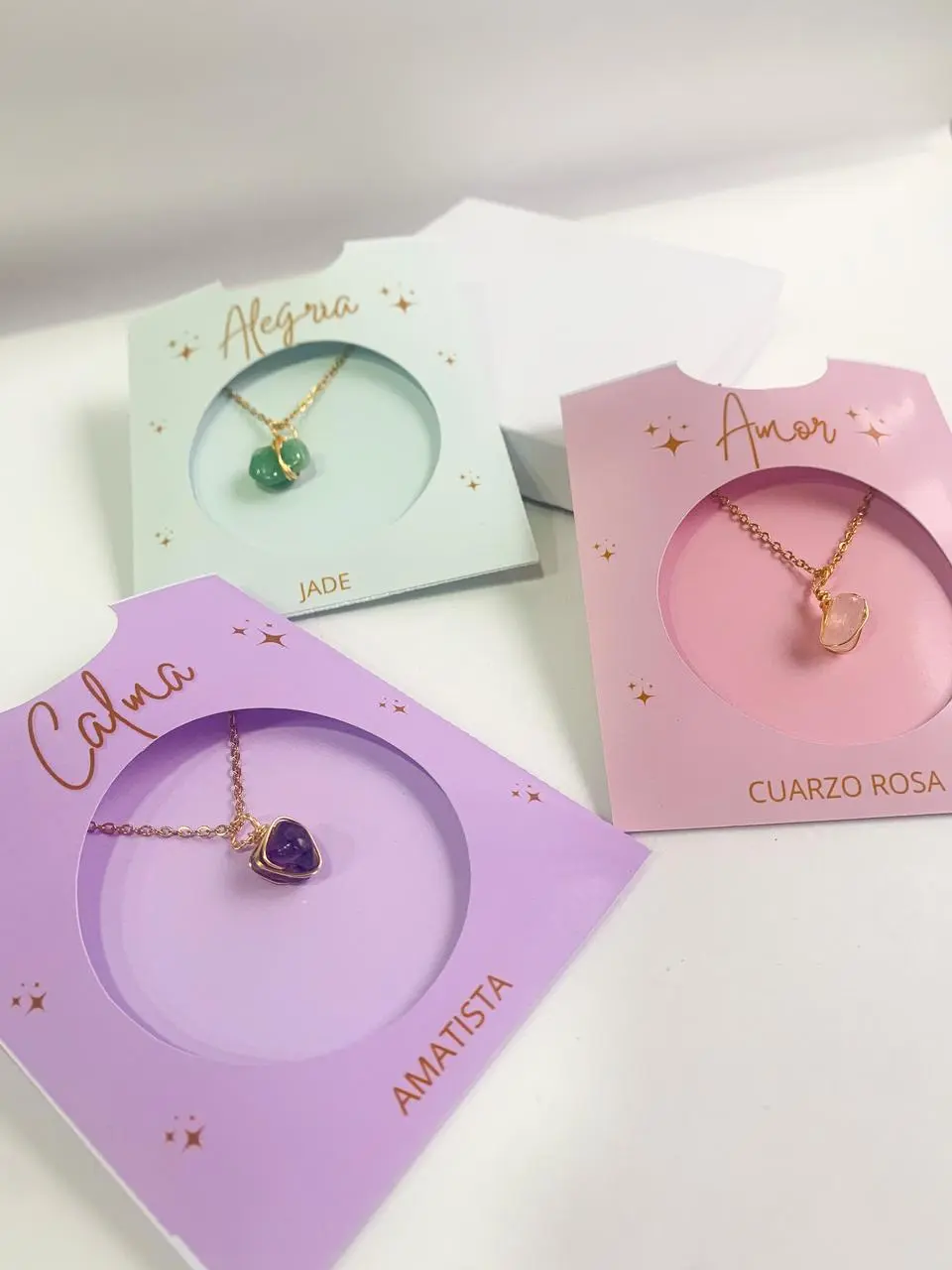 Collares artesanales de piedras naturales con dije envuelto en alambre dorado sobre tarjetas de presentación en tonos pastel: verde, lila y rosa.