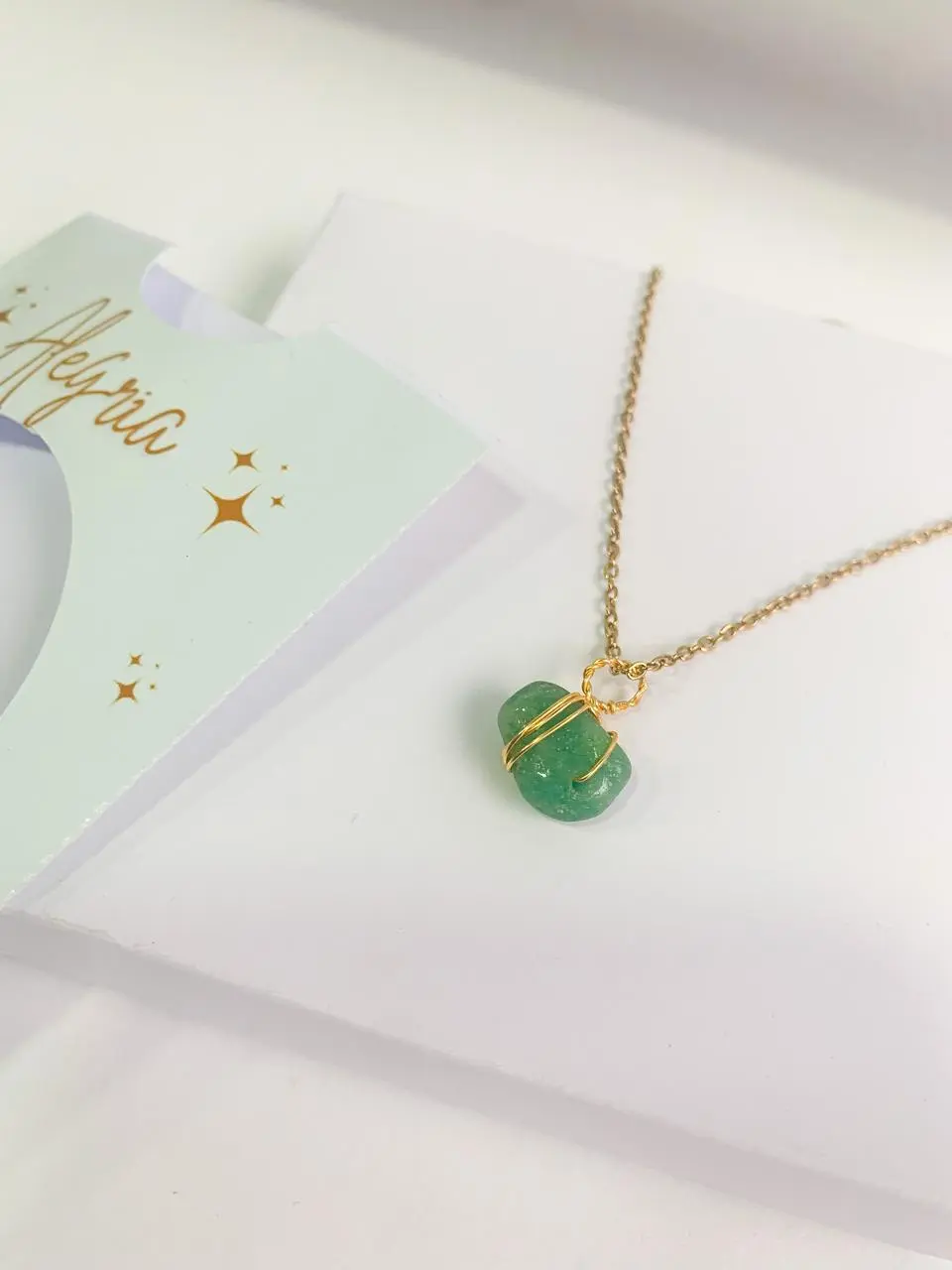 Collar artesanal con dije de piedra verde envuelto en alambre dorado y cadena fina, sobre empaque de marca en tonos menta.