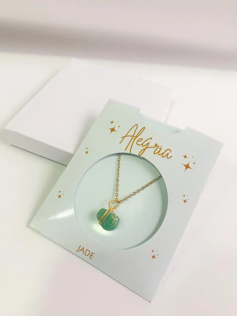 Collar con dije de jade verde envuelto en alambre dorado, sobre empaque claro con detalles dorados y caja blanca.