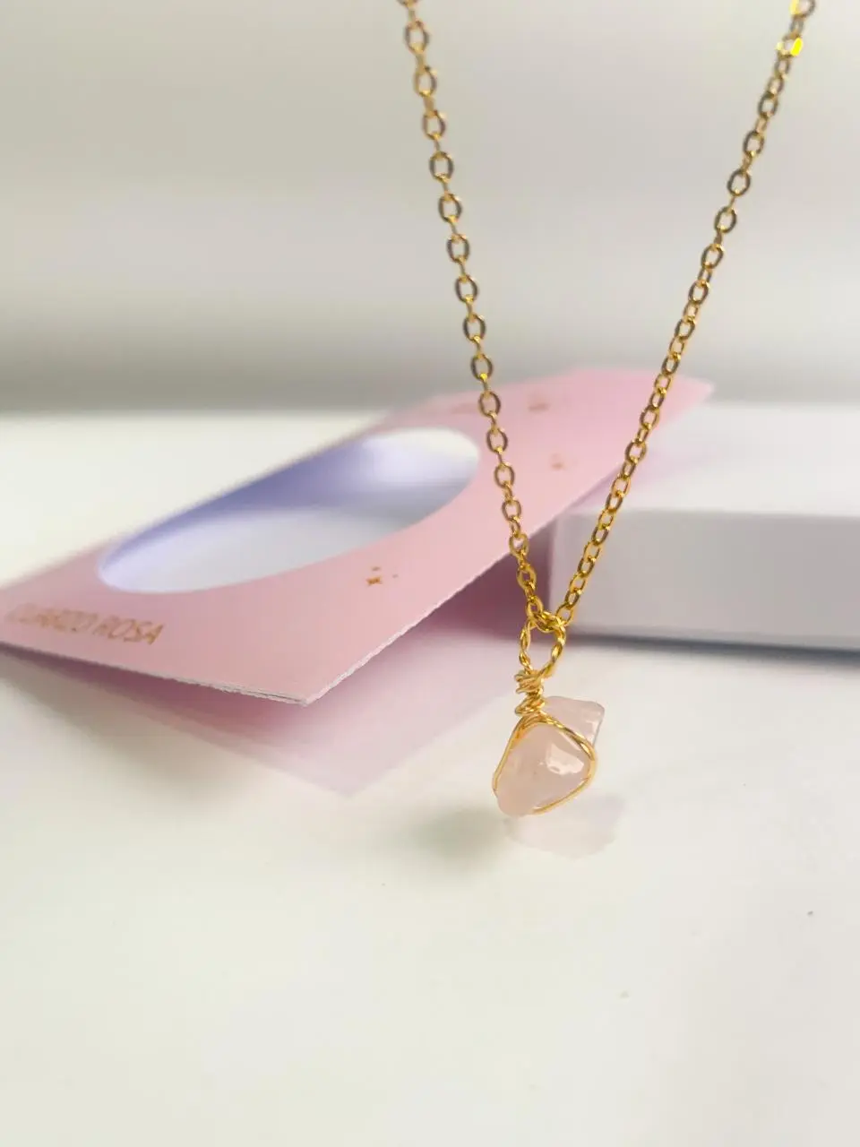 Collar artesanal de cuarzo rosa con dije de piedra natural envuelto en alambre dorado y cadena eslabonada, sobre fondo blanco y empaque rosa.