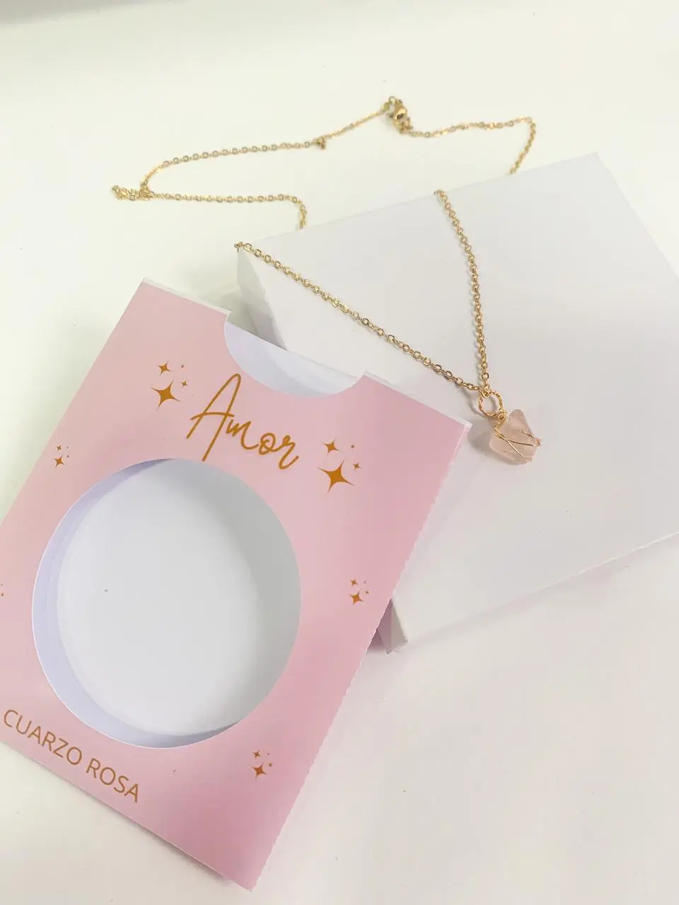 Collar de cuarzo rosa con dije de piedra natural envuelta en alambre dorado, sobre caja blanca y empaque rosado con la palabra Amor.