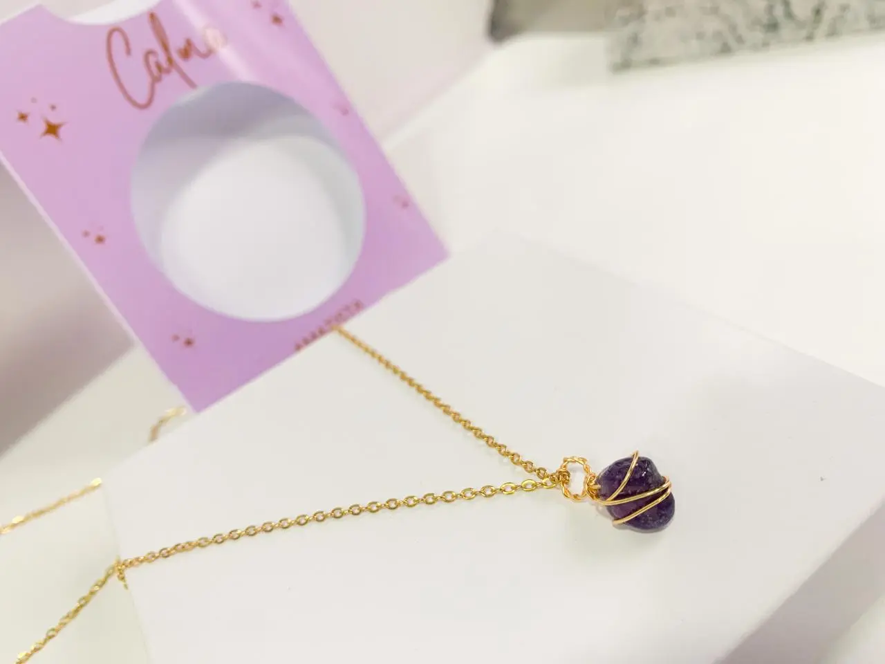 Collar dorado con dije de piedra natural amatista morada envuelta en alambre, sobre caja blanca y empaque lila.