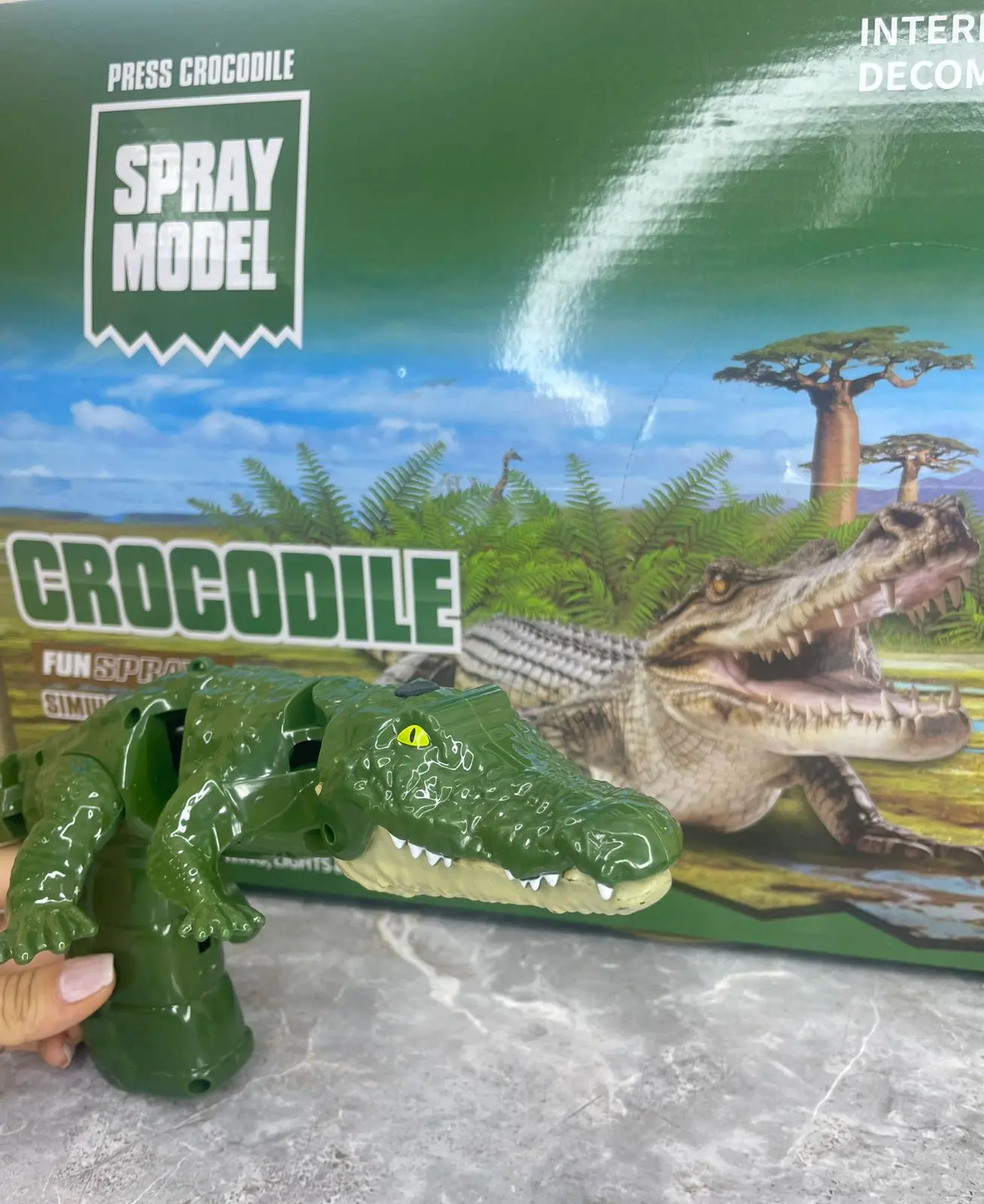 Pistola de juguete con forma de cocodrilo verde brillante, articulada y con efectos de spray, sosteniéndose sobre fondo de caja con diseño selvático.
